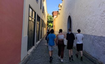 Eine Gruppe von vier Personen geht eine schmale, gepflasterte Straße entlang, umgeben von bunten Gebäuden und blauem Himmel.