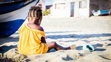 Ein kleines Kind sitzt im Sand, schaut auf einen Strand, mit einem Boot im Hintergrund und einem leuchtend gelben T-Shirt.