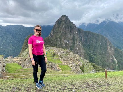 Eine Frau in einem pinken T-Shirt steht vor der beeindruckenden Kulisse von Machu Picchu, umgeben von grünen Bergen.