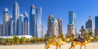 Zwei Kamele mit einem Reiter ziehen durch den Sand, während die beeindruckende Skyline von Dubai im Hintergrund leuchtet.