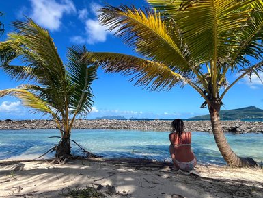 Eine Person sitzt am Strand, umgeben von Palmen, und blickt auf das ruhige Wasser und die Berge im Hintergrund.