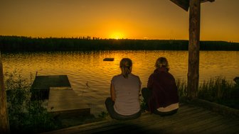 Zwei Jugendliche sitzen am Ufer eines Sees und beobachten den Sonnenuntergang, während die Wasseroberfläche golden schimmert.