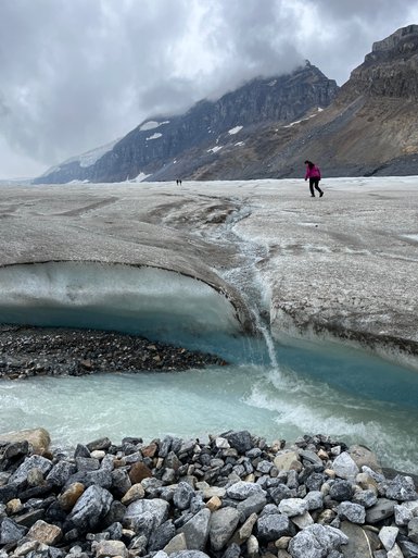 Eine Person in einem pinken Mantel wandert über das Athabasca-Eisfeld, umgeben von schroffen Bergen und grauem Himmel.