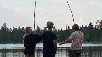 Drei Kinder stehen auf einem Steg am Wasser, halten lange Stöcke und blicken auf den ruhigen See im Hintergrund.