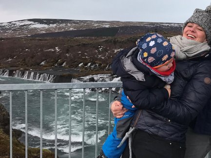 Eine lachende Frau hält ein kleines Kind in den Armen, während sie vor einem Wasserfall steht, umgeben von einer malerischen Landschaft.