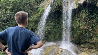 Ein Junge steht am Ufer eines klaren Wassers, während ein beeindruckender Wasserfall hinter ihm herabstürzt.