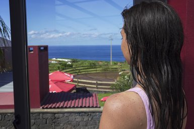 Eine Frau mit nassen Haaren blickt aus einem Fenster auf den Ozean und die grüne Landschaft im Hintergrund.