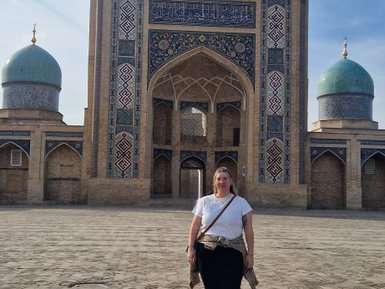 Eine Frau steht auf dem gepflasterten Platz vor dem beeindruckenden Hazrati Imam Komplex mit blauen Kuppeln im Hintergrund.