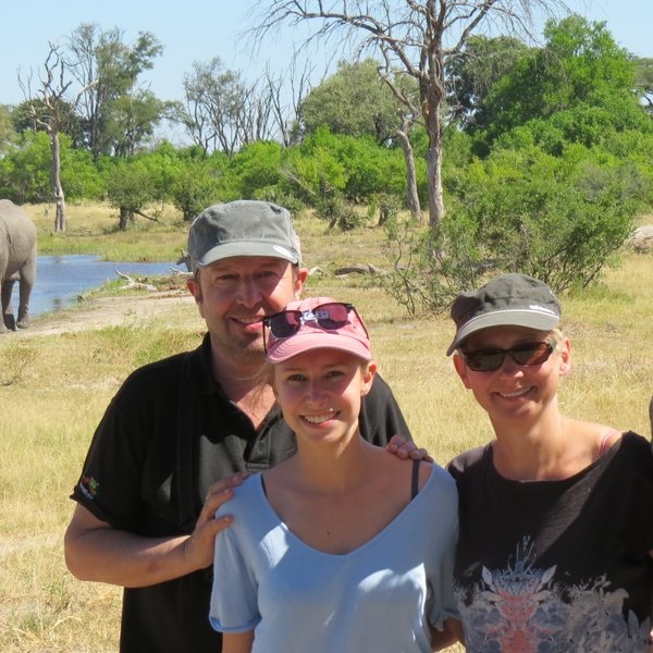 Eine Familie posiert fröhlich vor einem Elefanten, der im Hintergrund an einem Fluss steht, umgeben von grüner Natur.