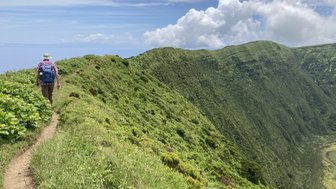 Ein Wanderer mit einem Rucksack geht einen schmalen Pfad entlang, umgeben von üppigem Grün und steilen Hängen.