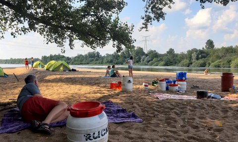 Am Strand entspannen Menschen, während Zelte im Hintergrund aufgestellt sind und der Fluss ruhig fließt.