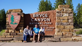 Eine Familie sitzt fröhlich vor dem Eingangsschild des Bryce Canyon Nationalparks, umgeben von Bäumen und klarem Himmel.