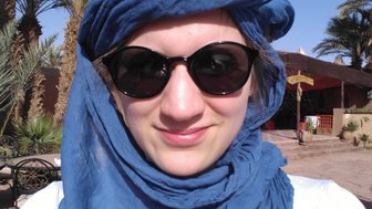 Eine Person mit einer blauen Kopfbedeckung und Sonnenbrille lächelt vor einer Palmenlandschaft und einem Gebäude.