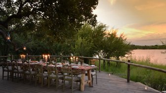 Ein festlich gedeckter Tisch mit Kerzen steht auf einer Holzterrasse, umgeben von üppigem Grün und einem ruhigen Gewässer.