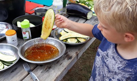 Ein Junge hält eine Zucchinihälfte über eine Schüssel mit Gewürzöl, während er am Tisch mit frischen Zutaten kocht.