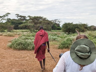 Ein Maasai in traditioneller Kleidung führt eine Person durch die savannenartige Landschaft Kenias.