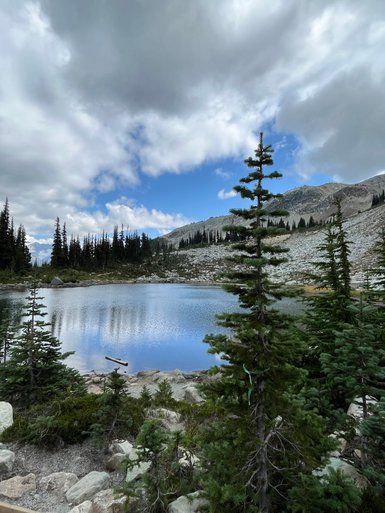 Ein ruhiger Bergsee spiegelt die Wolken und Bäume wider, umgeben von majestätischen Bergen und üppigem Grün.