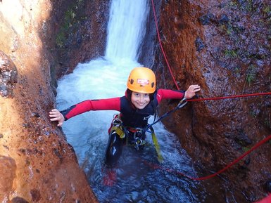 Ein fröhliches Kind in einem roten Neoprenanzug steht im Wasser, umgeben von steilen, moosbedeckten Felsen und einem Wasserfall.