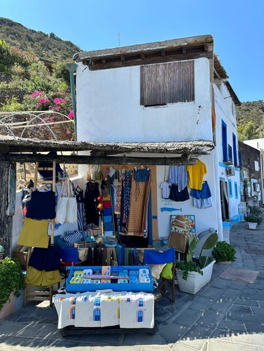 Ein charmantes Geschäft mit bunten Kleidungsstücken und Accessoires, umgeben von blühenden Pflanzen und einem klaren blauen Himmel.