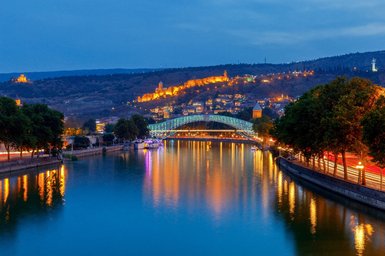 Die beleuchtete Brücke über den ruhigen Fluss spiegelt die Farben der Stadt Tiflis bei Nacht wider.