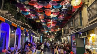 Bunte Regenschirme hängen über einer belebten Gasse, in der Menschen an Tischen sitzen und sich unterhalten.