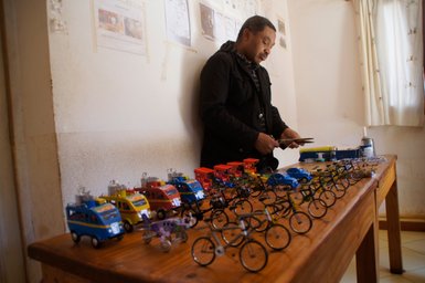 Ein Mann steht an einem Tisch, umgeben von bunten Spielzeugautos und Fahrrädern, während er mit einem Werkzeug arbeitet.