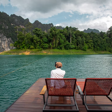 Ein Mann sitzt auf einer Holzterrasse und blickt auf das ruhige, grüne Wasser vor einer bewaldeten Berglandschaft.