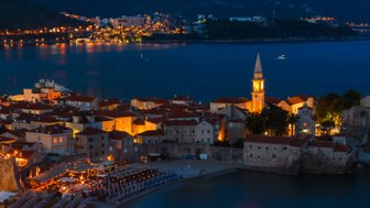 Die Küstenstadt Budva erstrahlt nachts in warmem Licht, während der Glockenturm im Vordergrund leuchtet.