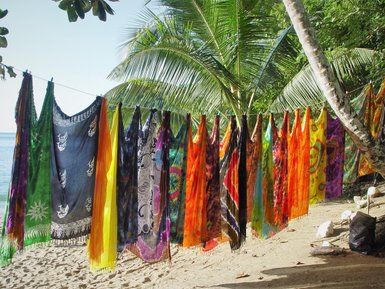 Bunte Tücher hängen an einer Leine, umgeben von Palmen und dem ruhigen Wasser eines Strandes im Hintergrund.