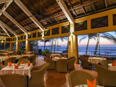 Ein elegantes Restaurant mit rattanartigen Stühlen, gedeckt mit weißen Tischdecken und orangefarbenen Servietten, bietet einen Blick auf den Sonnenuntergang.