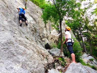 Ein Kletterer in blauer Kleidung erklimmt eine steile Felswand, während ein Sicherer mit Helm aufmerksam zusieht.