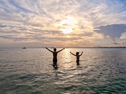 Zwei Personen stehen im Wasser und genießen den Sonnenuntergang, während sie fröhlich die Arme heben.