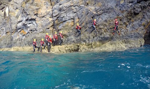 Eine Gruppe von Abenteurern in Neoprenanzügen klettert an einer steilen Felswand entlang, umgeben von klarem, blauem Wasser.