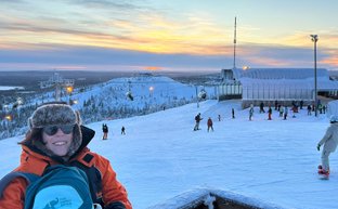 Eine Person mit einer orangefarbenen Jacke und einer blauen Tasche lächelt vor einer verschneiten Skipiste bei Sonnenuntergang.
