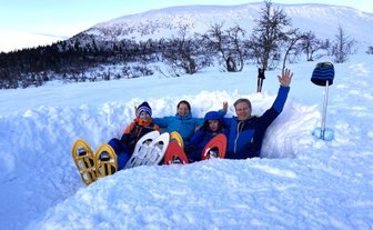 Eine Familie sitzt im Schnee, umgeben von schneebedeckten Bergen, und winkt fröhlich in die Kamera.