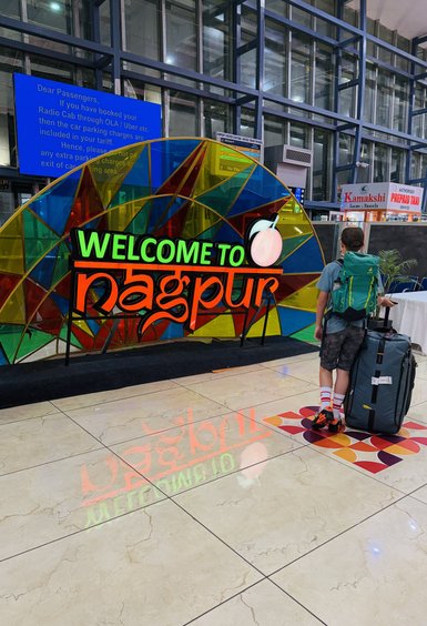 Ein Junge mit einem großen Koffer steht vor einem bunten Willkommensschild für Nagpur am Flughafen.