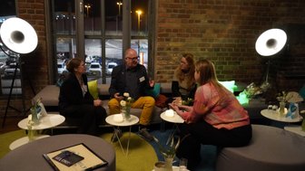 Vier Personen sitzen in einem gemütlichen Raum, umgeben von Pflanzen und sanftem Licht, während sie angeregt diskutieren.