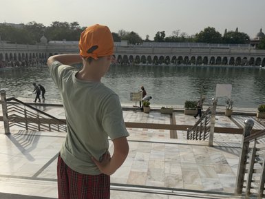 Ein Junge mit einem orangefarbenen Turban steht am Ufer eines ruhigen Gewässers und blickt auf die Menschenmenge.