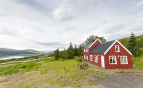 Ein rotes Sommerhaus steht malerisch in einer grünen Landschaft mit Blick auf einen ruhigen See und sanfte Hügel im Hintergrund.