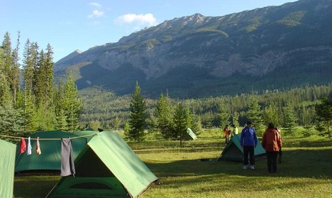 Zwei Personen stehen auf einem Campingplatz mit grünen Zelten, umgeben von hohen Bäumen und majestätischen Bergen im Hintergrund.