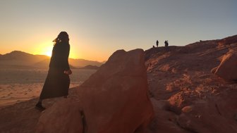 Eine Person in schwarzer Kleidung wandert über felsiges Terrain, während die Sonne hinter den Bergen untergeht.