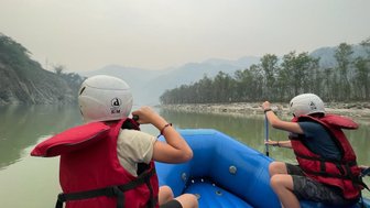 Zwei Kinder in roten Schwimmwesten paddeln in einem blauen Schlauchboot auf einem ruhigen Fluss in Nepal.