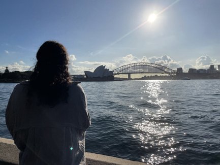 Eine Frau mit lockigem Haar blickt auf das Wasser, während die Sydney Opera House und die Harbour Bridge im Hintergrund sichtbar sind.