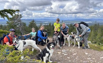 Eine Gruppe von Menschen posiert fröhlich mit mehreren Huskys auf einem Berg, umgeben von einer malerischen Landschaft.