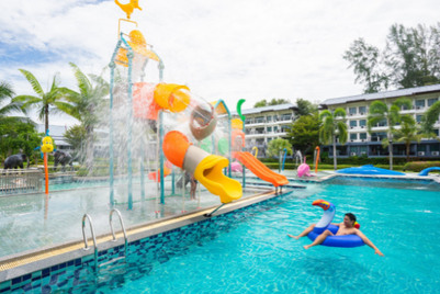 Ein Kind schwimmt entspannt im Pool, während ein Wasserspielplatz mit bunten Rutschen im Hintergrund spritzt.