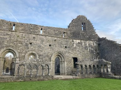 Die Ruine eines alten Steingebäudes mit gotischen Fenstern und Bögen, umgeben von grünem Gras und einem klaren Himmel.