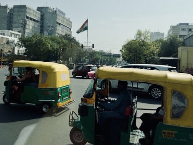 Zwei bunte Tuk-Tuks fahren an einer belebten Kreuzung in Delhi vorbei, während die indische Flagge im Hintergrund weht.