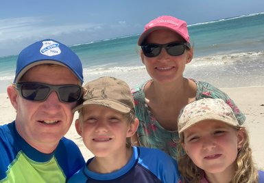 Eine Familie posiert am Strand, umgeben von türkisfarbenem Wasser und einem klaren Himmel, alle lächeln glücklich.
