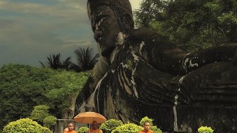 Drei Mönche in orangefarbener Robe gehen mit einem Regenschirm an einer großen Buddha-Statue vorbei, umgeben von üppigem Grün.
