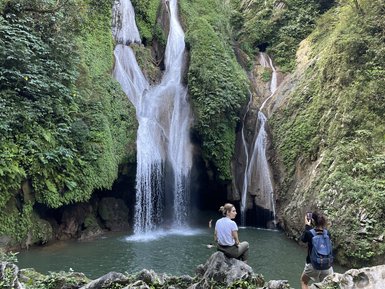 Ein Wasserfall stürzt in einen klaren Pool, umgeben von üppigem Grün und Felsen, während zwei Personen die Szenerie genießen.